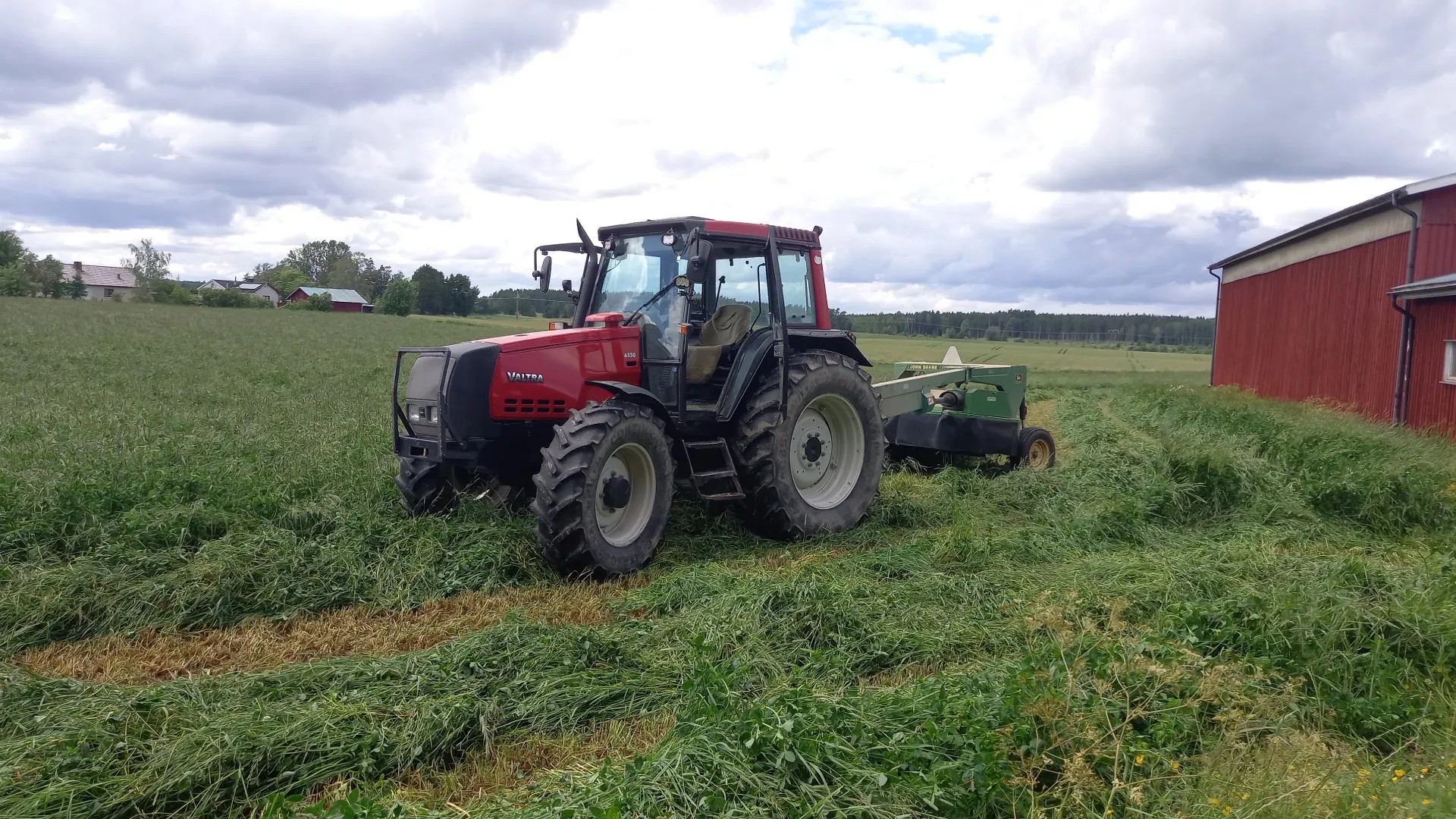 Röd traktor med slåtteraggregat på en nyslagen vall bredvid en röd ladugård